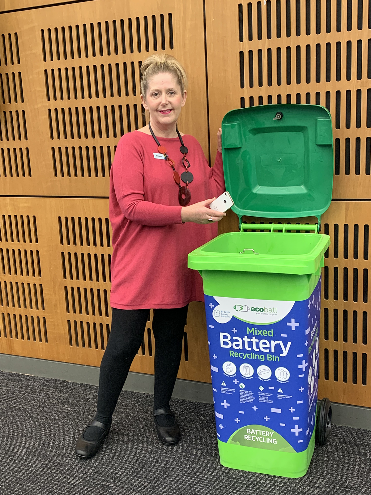 Woman placing an old mobile phone into a green battery and mobile phone recycling bin.